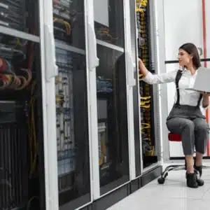 Technician checks equipment inside datacenter racks while referencing laptop, ensuring reliability and compliance procedures adherence