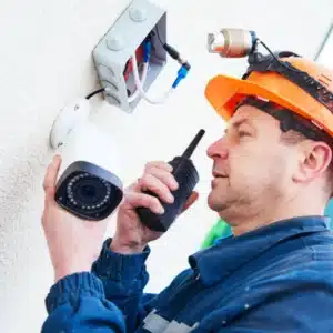 Technician with helmet and radio installs an outdoor wall-mounted security camera near exposed wiring