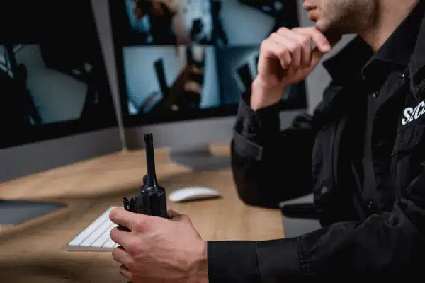 Security officer holds radio, monitors multiple surveillance screens, watching for incidents and coordinating responses tonight